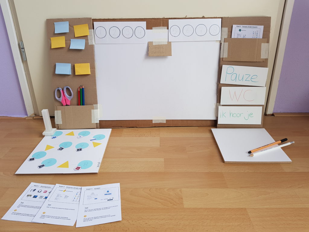 A low fidelity prototype made based on the paper prototype. A large rectangular piece of cardboard is propped upright. It has a blank area in the centre, about the size of a laptop screen. Above and to the sides there is space on the cardboard. Above the screen two sets of four round markers are visible. On the left there is a small pencil-bin and space above it to stick notes. On the right there a card holder with cards in it. Below the card holder there are three signs: one with the writing "Break", another marked "Bathroom break" and the last one marked "I can hear you".  In front of the board, on the left side, there is a board with blue round markers and yellow triangular markers, along with a pawn. On the right side, in front of the board, a mini-whiteboard with writing tools. 