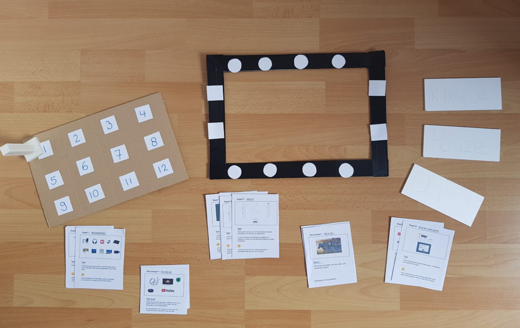 A photo of the paper prototype created by Denise Wien. This early prototype shows a cardboard rectangle with 12 numbered paper squares, a pawn, a black rectangular edge, oriented in landscape, with four round buttons on each long edge, and two square ones on each short edge. Below the two boards are five sets of cards. On the right three white rectangles. 